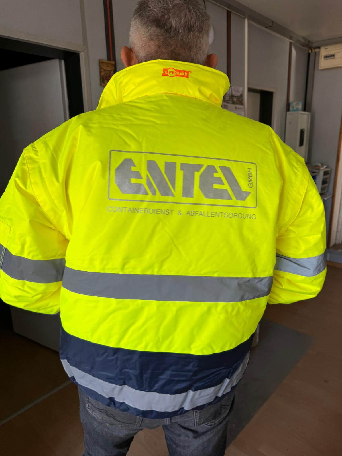 Entel-Mitarbeiter von hinten in gelber Warnschutzjacke mit Firmenlogo "Containerdienst & Abfallentsorgung"