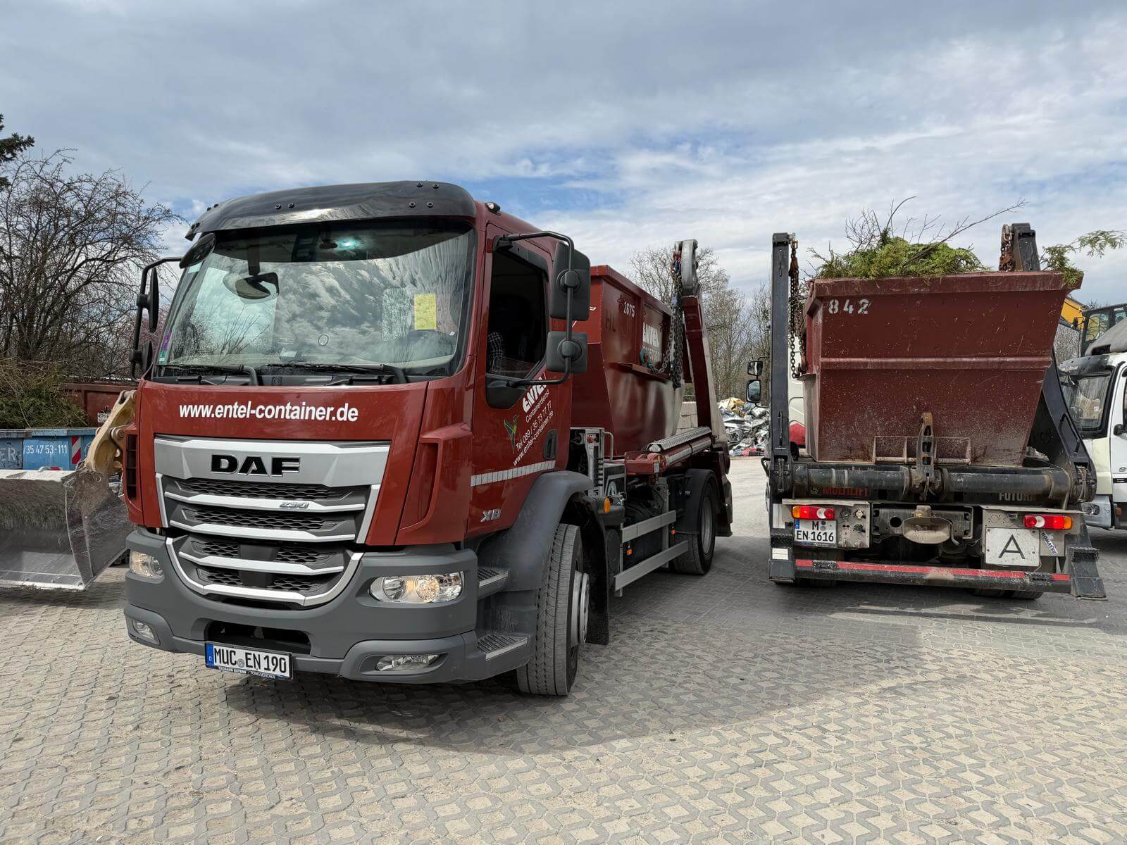 Roter Entel-LKW mit Absetzcontainer neben einem Anhänger mit Grünschnitt-Container auf dem Schrottplatz